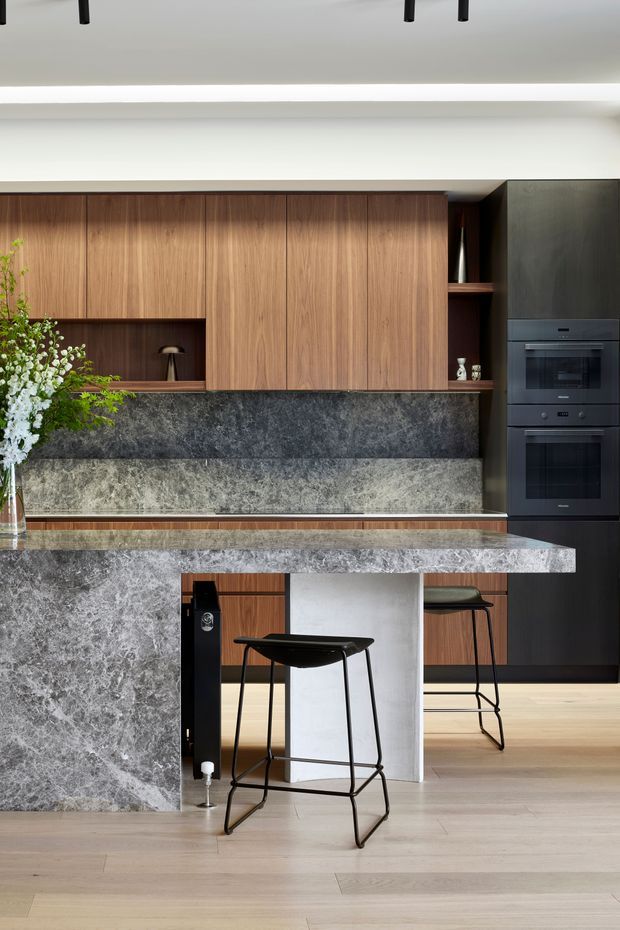 A contemporary kitchen with a large stone island and integrated waterfall-style side panel, warm timber cabinetry, dark built-in appliances, and a dark stone splashback. Black bar stools sit beneath the island, while open shelving and concealed linear ceiling lighting add refined detail. Pale timber flooring and a floral arrangement soften the otherwise high-contrast, sophisticated palette.