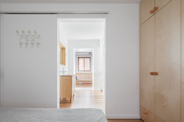 Bright minimalist bedroom viewed toward an open sliding doorway and hallway. The room features a pale grey wall, a low grey upholstered bed in the foreground, a full-height light timber built-in wardrobe with small rounded wooden pulls, and a simple wall-mounted coat rack. Beyond the doorway are light timber floors, white walls, pale timber cabinetry, and a distant window seat or radiator cover.
