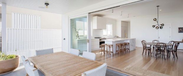 Bright open-plan interior featuring a dining area in the foreground, a white kitchen with island and bar seating beyond, and a second dining area to the right. The space has pale timber flooring, white walls and ceilings, large glazed openings, vertical white screening, and contemporary pendant lighting.