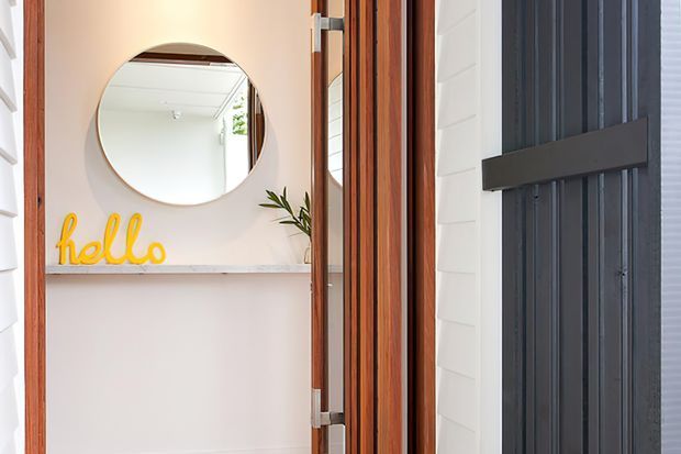 A bright residential entryway viewed from the exterior, featuring a timber-framed open door, white weatherboard cladding, and a dark vertical-panel front door with a horizontal handle. Inside the entry, a round wall mirror, pale stone or marble-look shelf, small plant, and yellow “hello” sign create a welcoming detail. The scene has a clean, coastal-inspired appearance with warm timber accents.