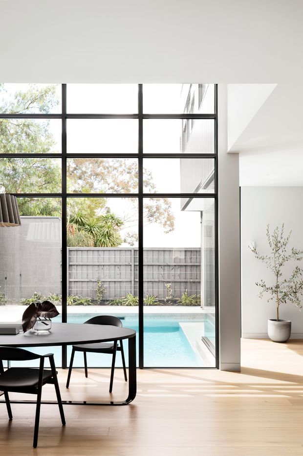 Bright contemporary dining area with a black-framed, floor-to-ceiling glazed wall overlooking a private swimming pool and landscaped courtyard. The room features pale timber flooring, white walls, a minimalist dark dining table and chairs, and a potted indoor tree. Strong natural light and clean geometric lines create an airy indoor-outdoor connection.