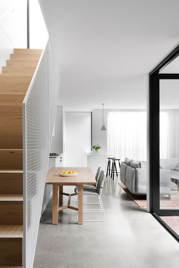 Bright minimalist open-plan interior viewed from beside a timber staircase. A light wood dining table with chairs sits in the foreground, opening into a white kitchen and living area with pale furnishings, sheer curtains, polished concrete flooring, and large black-framed glazing. A white mesh stair balustrade and clean-lined pendant light reinforce the contemporary aesthetic.