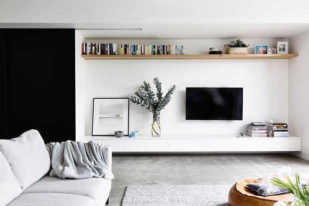 Bright, contemporary living room with a low white built-in media console, wall-mounted television, long timber display shelf with books and decor, and a large black feature wall. A pale sectional sofa, soft grey rug, coffee table and greenery create a clean, refined interior.