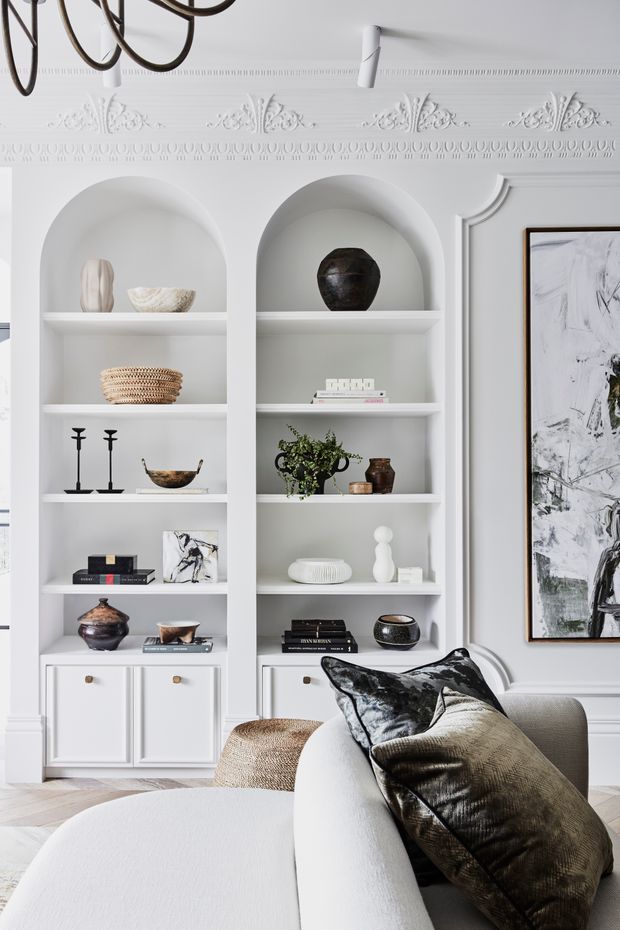 A refined living room with built-in white arched display shelving, decorative moulding and panelled detailing. The shelves contain curated ceramics, books, woven baskets and objects, while a large framed artwork and a pale upholstered sofa with textured cushions occupy the foreground. The palette is predominantly white and off-white with black and dark brown accents, creating a bright, elegant, traditional-meets-contemporary interior.