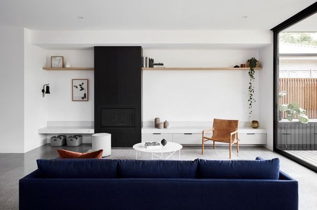 Bright contemporary living room with a deep navy sectional sofa in the foreground, a minimalist white built-in media and storage wall, a tall black fireplace surround, open timber shelving, a white sculptural coffee table, and a timber lounge chair. Full-height glazing opens to an exterior balcony or courtyard, bringing in abundant natural light. The room has a restrained, refined palette of white, black, navy, timber, and grey.