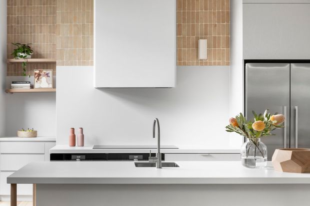 Bright contemporary kitchen featuring white flat-panel cabinetry, a large pale engineered-stone island benchtop, integrated black appliances, a stainless-steel double-door refrigerator, and a minimalist undermount sink with gooseneck tap. Warm beige vertical stacked tile forms a backsplash accent around open timber shelving, while a central white rangehood is set against a clean white wall.