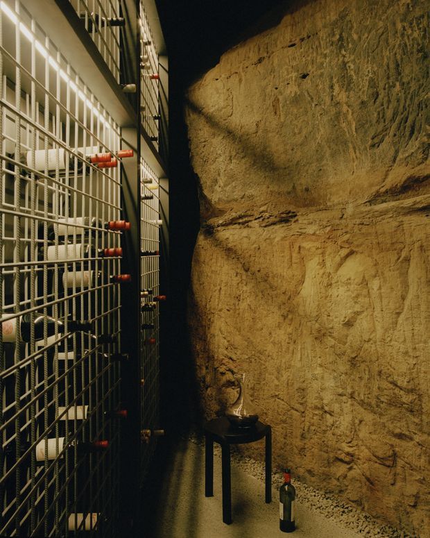A dimly lit wine storage space with floor-to-ceiling metal bottle racks on the left and an exposed rough ochre-brown stone or earth wall on the right. Bottles are stored horizontally in the gridded racks, with a small dark table and bottle in the narrow central passage. The atmosphere is rustic, intimate, and cellar-like.