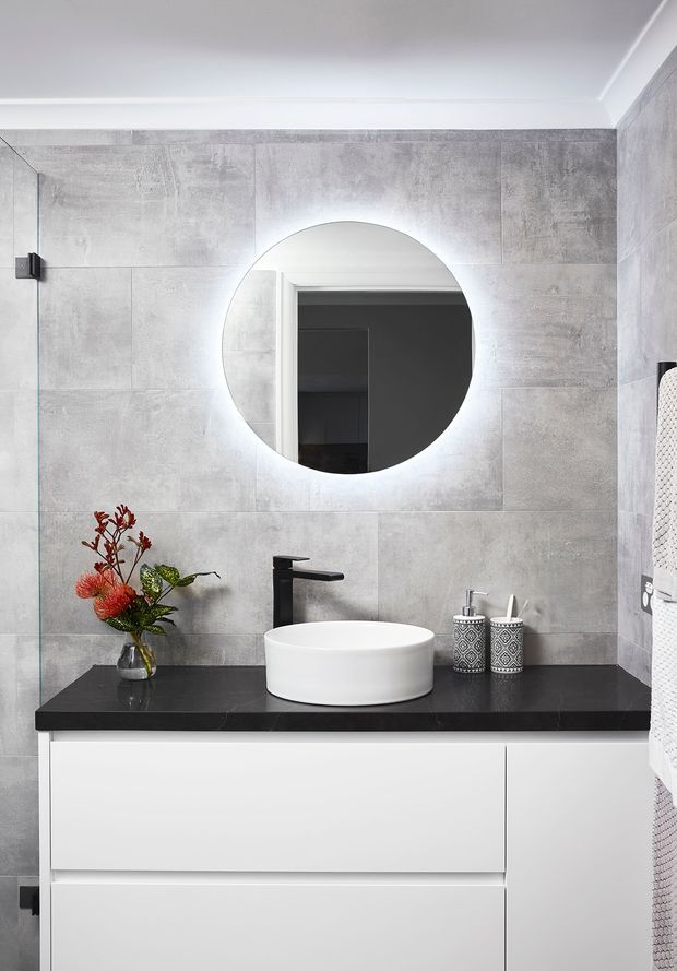 A contemporary bathroom vanity with a round backlit mirror, white vessel basin, matte black tapware, black stone-look benchtop, and white handleless cabinetry. Large grey concrete-look wall tiles create a clean, modern finish, with a small floral arrangement and patterned bathroom accessories adding contrast.