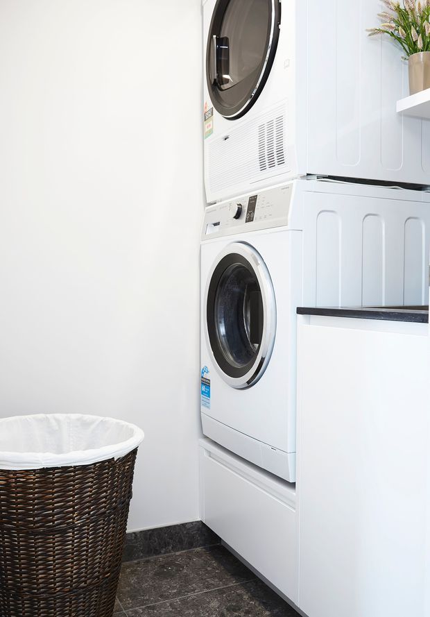 A compact, bright laundry room with a stacked white front-loading washer and dryer integrated into white cabinetry. The room has dark stone-look floor tiles, a dark benchtop, white paneled cabinetry, a wall shelf with a vase of foliage, and a woven laundry hamper.