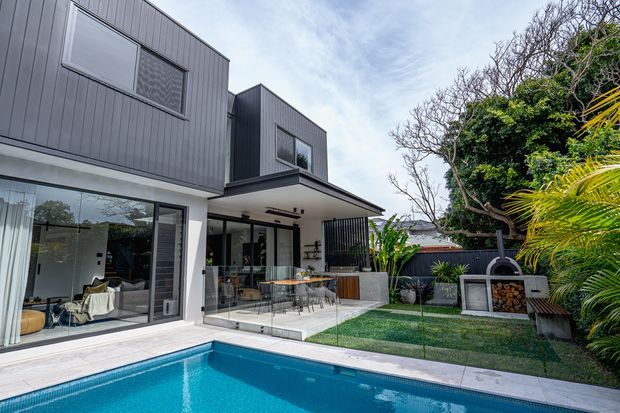 Contemporary two-storey home with dark vertical cladding and extensive glazing, opening onto a covered outdoor entertaining area and a bright blue swimming pool. The landscaped backyard includes a built-in outdoor fireplace or pizza oven and tropical planting.