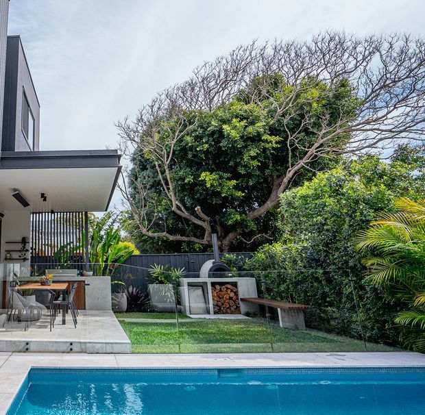 A contemporary residential backyard with a turquoise swimming pool in the foreground, a paved outdoor dining and entertaining area, and a built-in outdoor fireplace or pizza oven set into a landscaped garden. The house features dark vertical screening, large glazing, a flat roof, and clean concrete finishes, with mature tropical and subtropical planting surrounding the space.