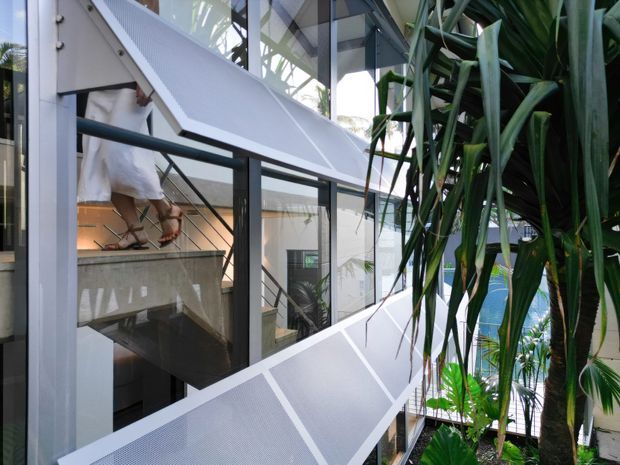 A contemporary multi-level interior atrium or circulation space with extensive floor-to-ceiling glazing, dark metal framing, angular perforated metal stair or walkway elements, exposed structural detailing, and abundant indoor tropical planting. A person is visible on a concrete stair, with additional levels and greenery seen through the glass.