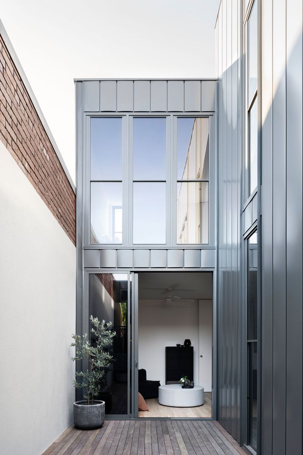 A narrow urban courtyard or passage leading to a contemporary home extension. The space features a timber deck, tall full-height glazing, dark grey metal cladding and framing, a white rendered wall with exposed brickwork above, and a partially open glazed door revealing a minimalist living room. The vertical proportions, reflective windows and restrained palette create a bright, modern architectural composition.