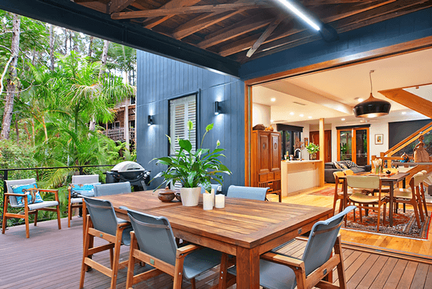 A spacious covered outdoor dining deck with a large solid timber dining table and upholstered timber-framed chairs, opening directly into a warm contemporary interior. The deck has dark blue vertical cladding, exposed timber roof framing, integrated lighting, lush tropical planting, and an adjacent living and kitchen area visible through wide openings. The interior includes timber cabinetry, a dining area, patterned rug, and staircase.
