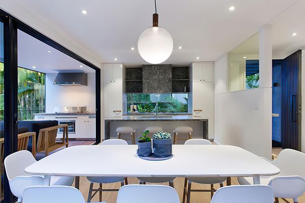 Bright open-plan dining and kitchen interior with a large white dining table and contemporary chairs in the foreground. The kitchen features white cabinetry, a dark textured stone backsplash, integrated appliances, a central sink beneath a window, timber flooring, black-framed glazing, and a sculptural pendant light. A living area is visible beyond the open partition.
