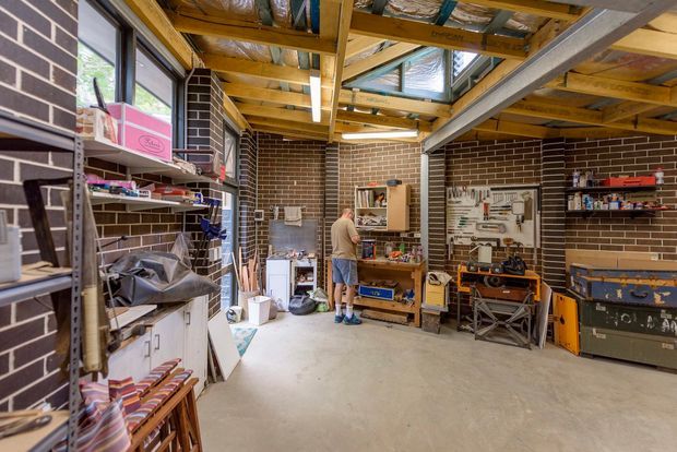 A spacious workshop or garage with exposed timber roof framing, skylights, dark brown brick walls, concrete flooring, open metal shelving, workbenches, tools, storage cabinets, and a large work area. The room has an industrial, practical character and abundant natural light.