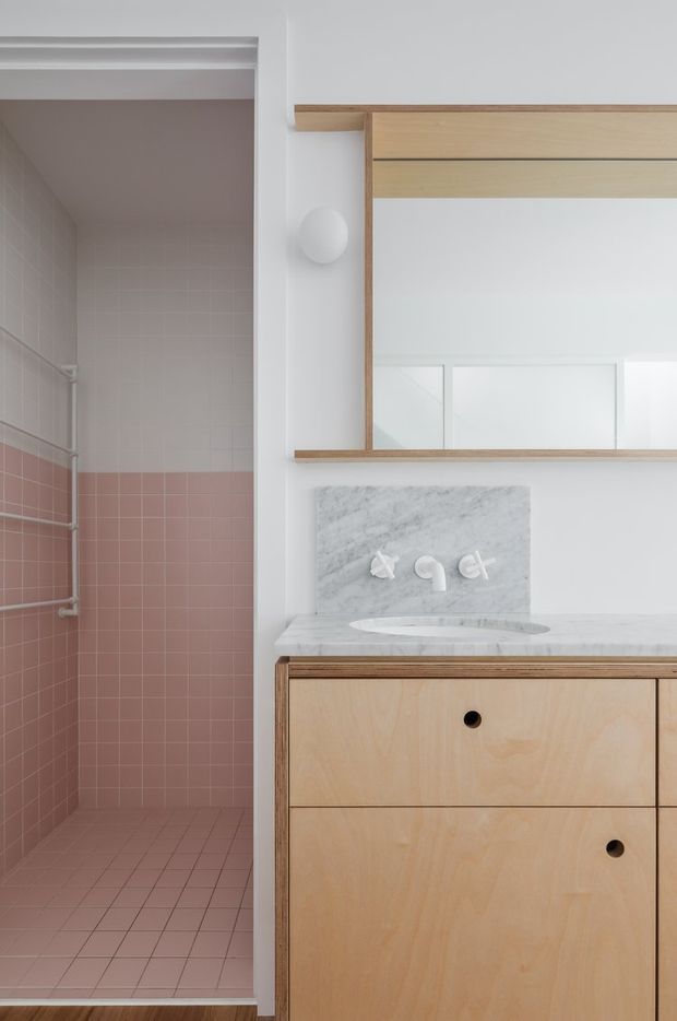 A clean, contemporary bathroom with a walk-in shower clad in pale pink square tiles, a floating or built-in light timber vanity, white marble benchtop and splashback, integrated oval basin, brushed light-coloured fixtures, and a large mirrored cabinet framed in timber. White walls and simple globe wall lighting create a soft, refined atmosphere.
