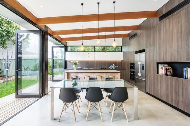 Bright contemporary open-plan kitchen and dining area with a large glass dining table, black dining chairs, timber cabinetry, a long island with bar stools, integrated appliances, and exposed timber ceiling beams. Full-height glazed sliding doors open to an outdoor garden area, creating strong indoor-outdoor connection. Pendant lights and warm timber finishes add contrast to the light neutral surfaces.