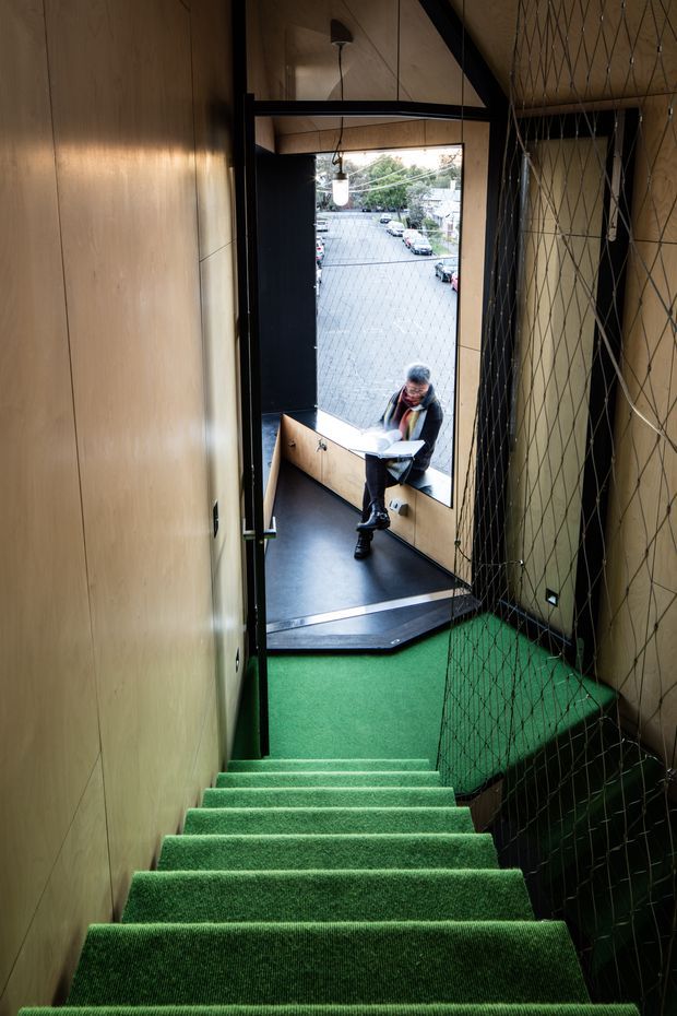 A narrow, steep interior stairway descends toward a small landing with a large open doorway overlooking a paved urban street. The stairs and landing are covered in vivid green carpet, while the walls and ceiling are clad in warm timber panels with dark metal framing. Protective netting runs along the right side, and a person sits at a built-in timber counter near the entrance. The space has a compact, unusual, utilitarian and playful architectural character.