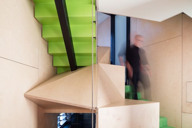 Interior view of a compact, highly contemporary stairway featuring vivid green stair treads and risers, pale timber or plywood wall and stair surfaces, and a bold dark structural handrail or beam. The image is composed around the stair circulation and includes a blurred person moving through the space, with glimpses of adjacent levels and dark-framed glazing.