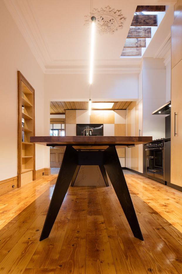 A warm, refined kitchen-dining space in a period-style interior. A large dark timber dining table with dramatic angled legs dominates the foreground, set over polished honey-toned timber flooring. Behind it is a contemporary kitchen with pale timber and white cabinetry, a dark stone or black splashback, integrated appliances, and a central island. The room features ornate ceiling cornices, a decorative plaster ceiling medallion, a timber-lined ceiling section, skylights with textured panels, and built-in timber shelving.