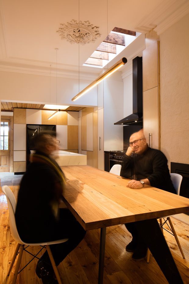 Warmly lit, narrow open-plan kitchen and dining area with a long solid timber dining table, contemporary built-in cabinetry, black rangehood and appliances, timber flooring, and a large roof skylight. The room combines ornate traditional ceiling mouldings and a decorative ceiling medallion with modern joinery and linear pendant lighting. Two seated people are present, with motion blur in the foreground.
