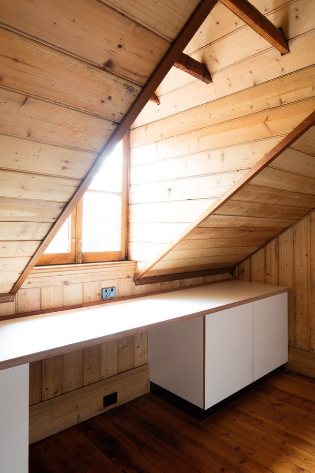 A compact attic-like interior with extensive warm timber lining on the sloped ceiling and walls, exposed dark timber rafters, a small timber-framed window, and a long white built-in desk or cabinet running beneath the window. The room has dark timber flooring and appears to function as a small workspace or study, though no chair or other office equipment is visible.