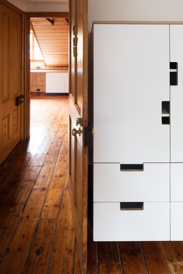 A narrow interior passage or room with polished warm timber floorboards, timber wall panelling, and a sloped timber-lined ceiling visible beyond an open traditional wooden door. A large minimalist white built-in storage unit with dark recessed handles occupies the right foreground, contrasting with the heritage timber detailing.