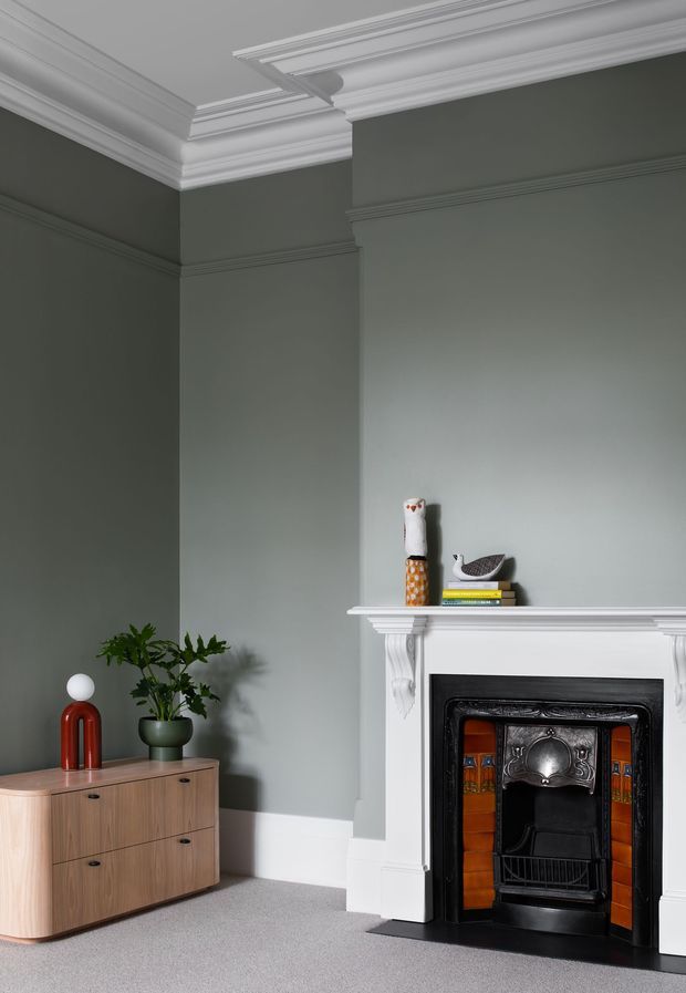 A refined living room corner featuring a dark sage-grey painted wall, ornate white decorative cornices and dado mouldings, and a restored fireplace with a white carved mantel, black cast-iron insert, and orange patterned tile surround. A low mid-century timber sideboard with rounded corners, a sculptural red table lamp, potted plant, and small decorative objects complete the composition. The room has a calm, elegant, heritage-inspired character with contemporary furnishings.