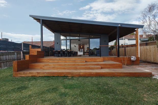 Rear exterior view of a contemporary single-storey backyard pavilion or extension with a broad low-pitched roof, dark framed glazing, exposed timber soffit, and a substantial tiered timber deck with integrated steps and seating. The sheltered indoor-outdoor entertaining area opens onto a lawn and is enclosed by timber fencing, with a dining setting visible inside.