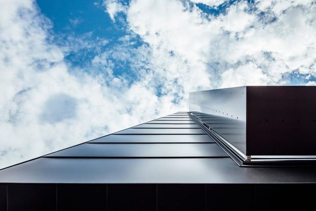Upward-looking close-up of a sleek contemporary building facade and roofline against a dramatic partly cloudy sky. The composition emphasizes smooth dark metal cladding, strong horizontal roof seams, and a projecting rectangular metal volume with crisp geometric detailing.