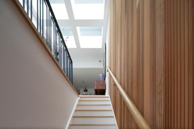 Bright contemporary stairway rising toward an upper landing, with pale timber stair treads and handrail, a vertical timber slat feature wall, dark metal balustrade, and a series of rectangular skylights or rooflights overhead. A small portion of the upper-level interior is visible beyond the stairs.