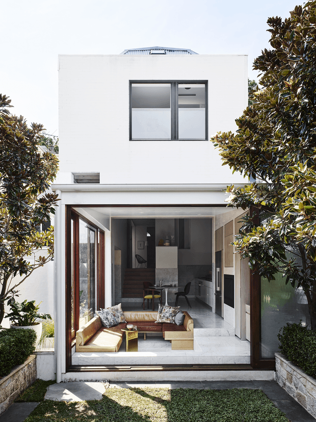 A bright view from a small landscaped courtyard into a contemporary two-storey home. Large timber-framed glazed doors open onto a compact living area with a tan sofa, patterned cushions, a low coffee table and armchair, while a kitchen and dining area sit beyond. The white rendered facade has a simple rectangular form, dark-framed upper window, flat roofline with a raised rooftop element, and greenery framing both sides of the entry.