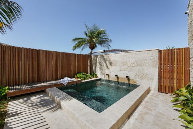 A private courtyard pool area with a compact rectangular swimming pool, pale stone surrounds, timber slatted privacy fencing, a textured masonry feature wall with multiple water spouts, built-in timber bench seating, and tropical palm planting under a clear blue sky.