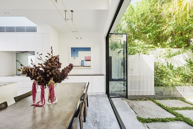 Bright contemporary open-plan kitchen and dining area with white full-height cabinetry, a large concrete-look dining table, black-framed glazing and a fully open glazed door leading to a lush landscaped courtyard. Minimal pendant lighting and polished concrete-style flooring create a clean indoor-outdoor connection.