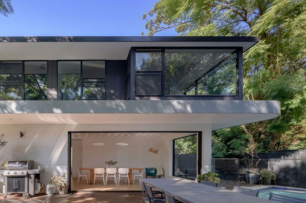 Modern two-storey residential exterior with a flat roof, dark cladding and extensive black-framed glazing. The lower level opens onto a timber deck and outdoor dining area, with a built-in barbecue and a swimming pool visible at the right. Mature trees surround the property.