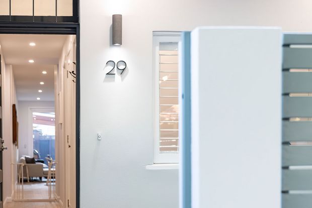 Bright contemporary residential hallway or entry area with pale walls, a narrow horizontal window with white plantation shutters, recessed ceiling lights, and a large pale built-in or partition element in the foreground. A dark-framed glazed transom and open doorway lead toward a furnished living or dining space. Minimal black wall numbers and a vertical wall light provide small graphic accents.