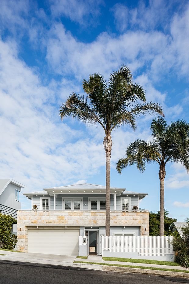 Two-storey coastal-style residence viewed from the street, with a pale stone-clad lower level, light grey upper-level weatherboard-style walls, broad verandah and louvred shutters. A double garage, white picket fencing, driveway and tall palm trees define the front elevation.