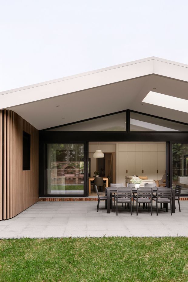 A contemporary single-storey house viewed from the garden, with a covered outdoor entertaining terrace and dining setting. The facade combines warm vertical timber screening with dark-framed floor-to-ceiling glazing, exposed brick at the base, and a broad flat roof with a rectangular skylight. The terrace is paved in large light-grey slabs and connects visually to an interior dining and kitchen area.