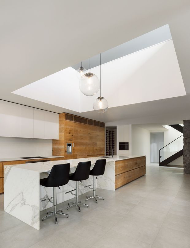 A bright, open-plan contemporary kitchen with a large white marble-look island and three black bar stools. The kitchen features sleek white handleless cabinetry, warm timber wall panelling and matching timber drawer fronts, integrated appliances, pale large-format flooring, and three pendant lights suspended beneath a dramatic angled ceiling opening. A hallway and stairway are visible beyond the kitchen.
