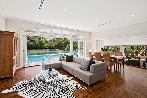 Bright, spacious open-plan living and dining area with polished dark timber flooring, white walls and ceiling, and extensive glazed bi-fold doors opening to a swimming pool and landscaped garden. The living zone contains a large neutral sectional sofa, glass coffee table, patterned rug, and timber cabinetry, while the adjacent dining area has a timber table with woven chairs. Classical wall and ceiling mouldings, French-style glazing, and white columns create a refined transitional interior.