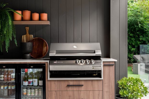 A compact outdoor kitchen or entertaining area featuring a built-in stainless-steel barbecue set into warm timber-look cabinetry, with a glass-front beverage refrigerator below the benchtop. The backdrop is dark vertical cladding, complemented by open shelving, terracotta pots, wooden boards, greenery, and a glass balustrade opening to a garden.