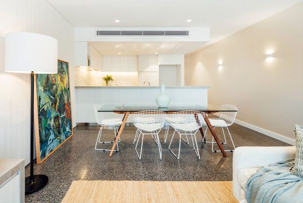 Bright open-plan dining and kitchen interior with a white dining table surrounded by wire-frame chairs, a polished speckled stone or terrazzo-like floor, pale cabinetry, and a light-toned kitchen island. The room has minimalist furnishings, recessed lighting, a large abstract artwork, and a contemporary neutral palette with warm timber accents.