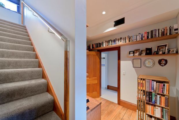 Interior view combining a carpeted staircase with timber handrail and trim on the left, and a warm timber-floored study or library on the right. The study features extensive built-in timber shelving filled with books, framed photographs, and displayed objects, plus a timber doorway leading to another room. Recessed ceiling lighting and warm natural materials create a traditional, inviting character.