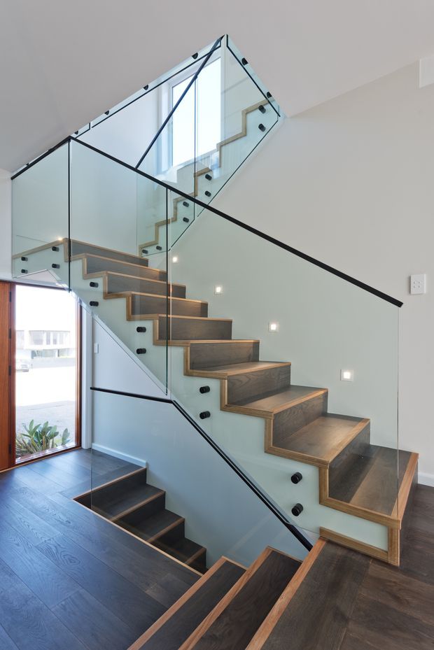 Interior architectural photograph focused on a modern stairway. The stair has dark timber treads with lighter timber edging and fully glazed balustrades secured with black fittings. A second flight and landing are visible through the glass, with recessed square wall lights and a tall window providing strong natural light. Dark timber flooring continues through the lower level.