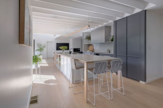 Bright, open-plan contemporary kitchen with a long pale island and integrated dining-height extension, white cabinetry, dark grey full-height storage, light timber flooring, and exposed white ceiling beams. The space is illuminated by extensive glazing or skylights and includes minimalist bar stools, pendant lighting, and restrained greenery.