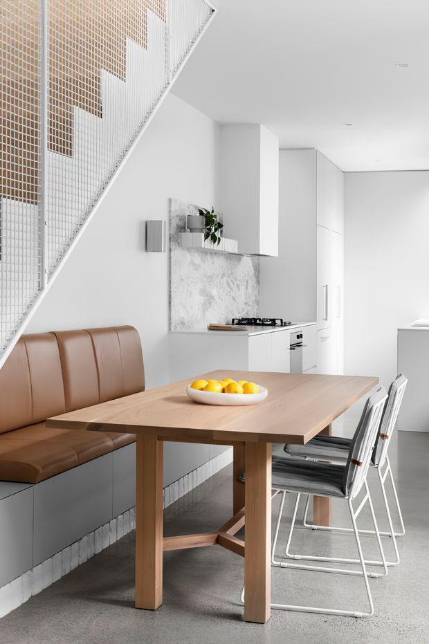 Bright contemporary open-plan dining and kitchen area beneath a staircase. A light timber dining table is paired with a built-in brown leather bench and folding metal-framed chairs. The compact white kitchen features minimalist cabinetry, a marble-look splashback, integrated appliances and a gas cooktop. White tiled stair detailing and a grey polished concrete floor complete the clean, refined interior.