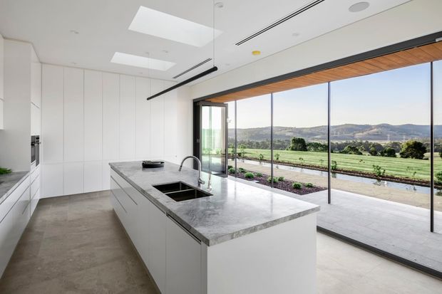 A bright, minimalist contemporary kitchen with a large white island, integrated sink and chrome tap, extensive floor-to-ceiling glazing, and white full-height cabinetry. A timber-lined ceiling overhang frames the glazed opening to a paved terrace and expansive rural landscape beyond. Skylights and linear recessed lighting enhance the airy, open interior.