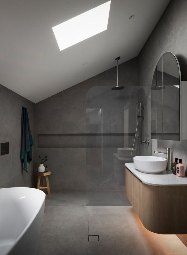 Contemporary bathroom with large-format grey wall and floor tiles, a freestanding white bathtub, walk-in glass shower, vessel sink, timber vanity, arched mirror, skylight, and subtle under-vanity lighting. The space has a refined, moody and minimalist character.