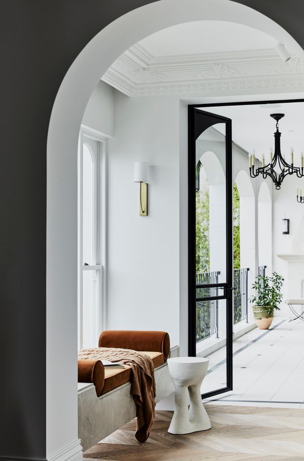 A refined white interior viewed through a large curved archway, featuring ornate decorative ceiling mouldings, white walls and trim, a black-framed glazed door, and a bright tiled veranda or courtyard beyond. The foreground includes a brown leather lounge chair, draped textile, sculptural white side table, herringbone timber flooring, classic wall lighting, and a black traditional chandelier. The space blends period architectural detailing with contemporary furnishings.